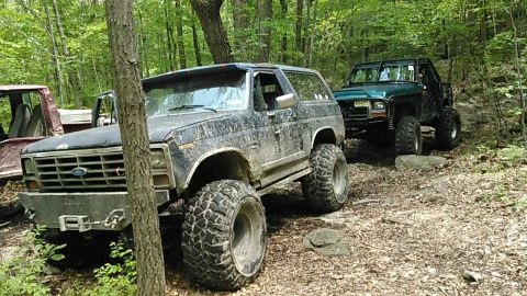 1988 Ford Bronco - Two modified Ford Broncos are parked on a wooded trail. The Bronco in the foreground is dark colored and appears to be a 1980s model, while…