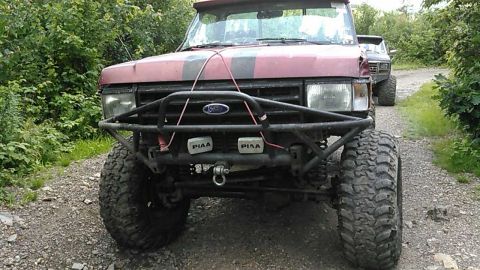 1988 Ford Bronco - The front view of a red Ford Bronco with a custom black bumper and large off-road tires is parked on a gravel road surrounded by trees. Two…