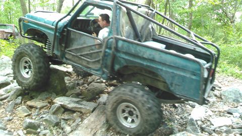 1988 Ford Bronco - A teal off-road modified pickup truck with a roll cage is navigating a rocky trail in a wooded area. The driver is visible inside the cab,…