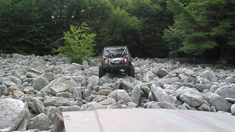 1988 Ford Bronco - A heavily modified off-road vehicle with a roll cage navigates a rocky terrain with a forest in the background.
