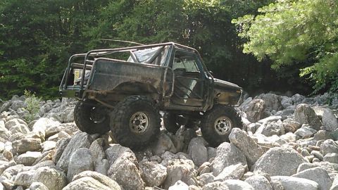 1988 Ford Bronco - A heavily modified, dark green off-road vehicle with a roll cage is navigating a rocky terrain with dense trees in the background.