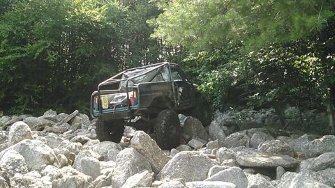 1988 Ford Bronco - A modified blue Ford Bronco with a roll cage navigates a rocky, wooded trail. The vehicle is positioned at an angle, with its front tires o…