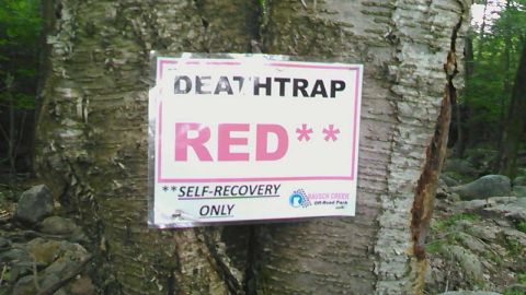 1988 Ford Bronco - A sign is attached to a tree in a wooded area. The sign reads "DEATHTRAP RED" and "SELF-RECOVERY ONLY" with a logo for "RAUSCH CREEK Off-Ro…