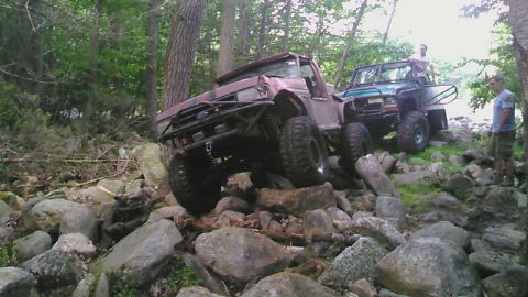 1988 Ford Bronco - A heavily modified, lifted red Ford Bronco is navigating a rocky, wooded trail, with a green Jeep-like vehicle and two men visible in the b…