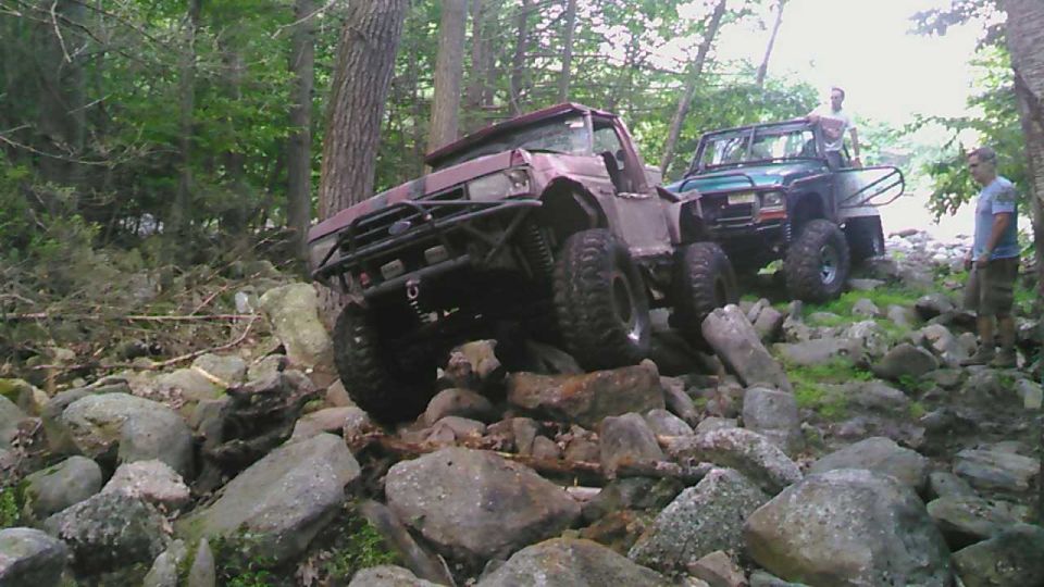 Modified red Ford Bronco off-roading on a rocky trail with a green Jeep and two men in the background.