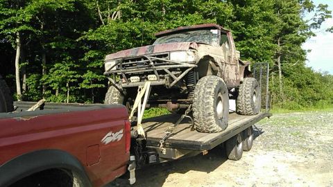 1988 Ford Bronco - A heavily modified, lifted off-road truck with large tires is loaded onto a flatbed trailer, which is being towed by a red pickup truck. Th…