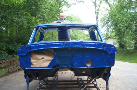 1979 Ford Bronco - A man is working on the roof of a bright blue vehicle body, which is mounted on a stand outdoors. The vehicle appears to be a truck or SUV…