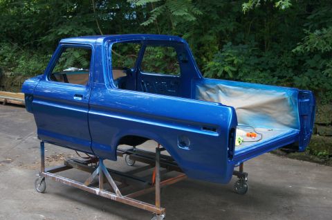 1979 Ford Bronco - A bright blue 1979 Ford Bronco body is shown mounted on a metal stand with wheels. The vehicle is outdoors with trees in the background, an…