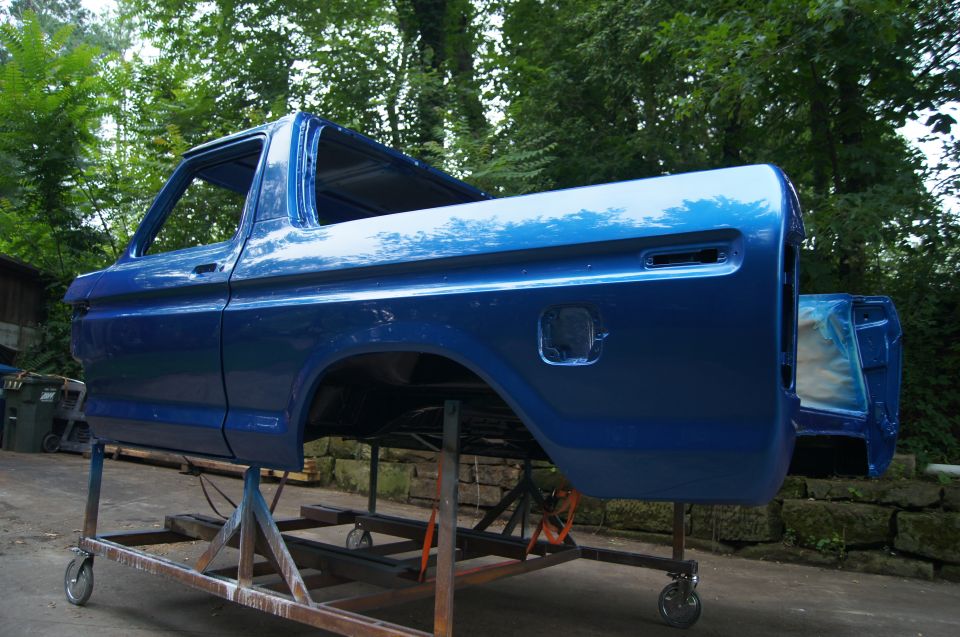 Blue classic pickup truck bed and cab on a rolling stand outdoors.