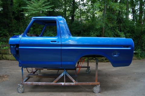 1979 Ford Bronco - The body of a blue 1979 Ford Bronco is shown on a rolling stand outdoors. The vehicle is missing its engine, interior, and front end, but t…
