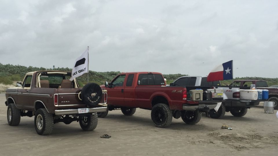 Brown 1978 Ford Bronco with spare tire on rear, next to red Ford F-Series truck on beach