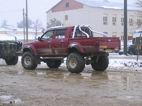1981 Ford Bronco - A lifted red pickup truck with a roll bar is seen in a muddy, snowy environment, next to another vehicle.