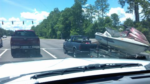 1994 Ford Bronco - A dark blue pickup truck towing a jet ski on a trailer drives down a road lined with trees. A red pickup truck is in the lane next to it.