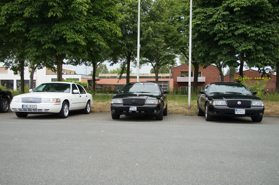 Three sedans parked in a row, one white and two black, with trees and buildings in the background.