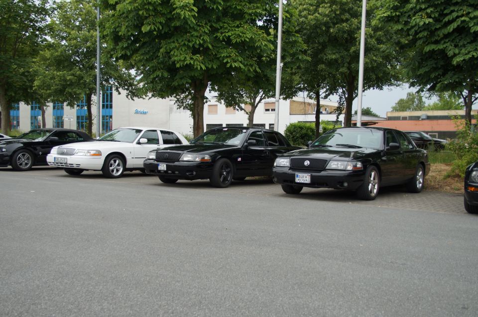 Four sedans parked in a row, three black and one white, with trees and a building in the background.
