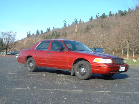 1999 Ford Crown Victoria - A red sedan is parked in an outdoor lot on a clear day with a wooded hill in the background.