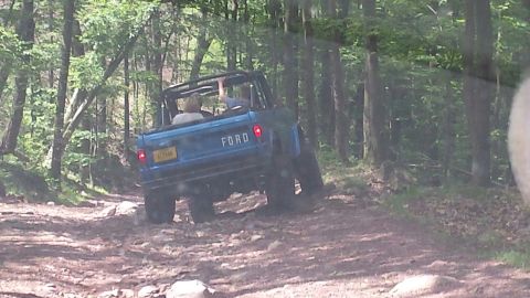 1974 Ford Bronco - A blue Ford Bronco drives on a rocky, wooded trail. The word "FORD" is visible on the tailgate.