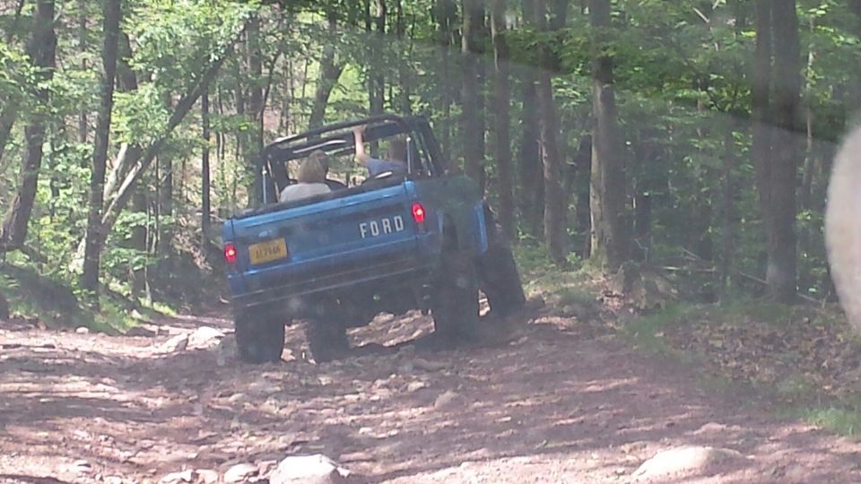 Blue 1974 Ford Bronco driving on a rocky forest trail.