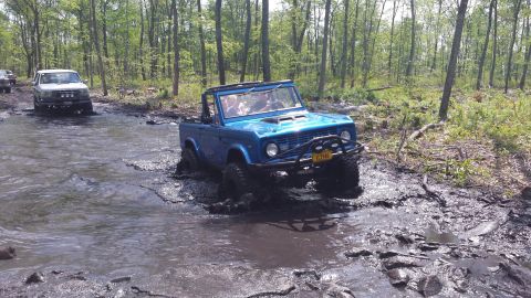 1974 Ford Bronco - A blue convertible Ford Bronco is driving through a muddy stream in a forest. Another white SUV is visible in the background also in the wa…