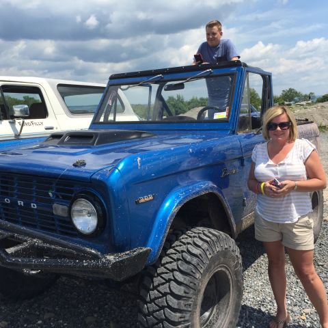 1974 Ford Bronco - A woman stands next to a lifted, bright blue Ford Bronco with large off-road tires. A man is visible in the driver's seat, leaning out of t…