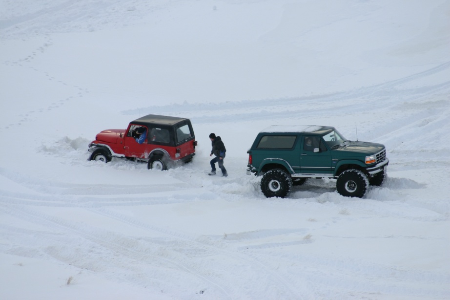 Green lifted Ford Bronco and red Jeep stuck in deep snow with a person standing between them