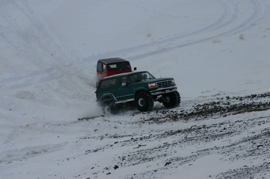 Green lifted Ford Bronco on oversized tires climbing snowy hill, red 4x4 behind