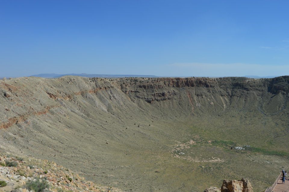 Wide view of a large impact crater with steep, rocky walls and a vegetated basin under a clear blue sky.