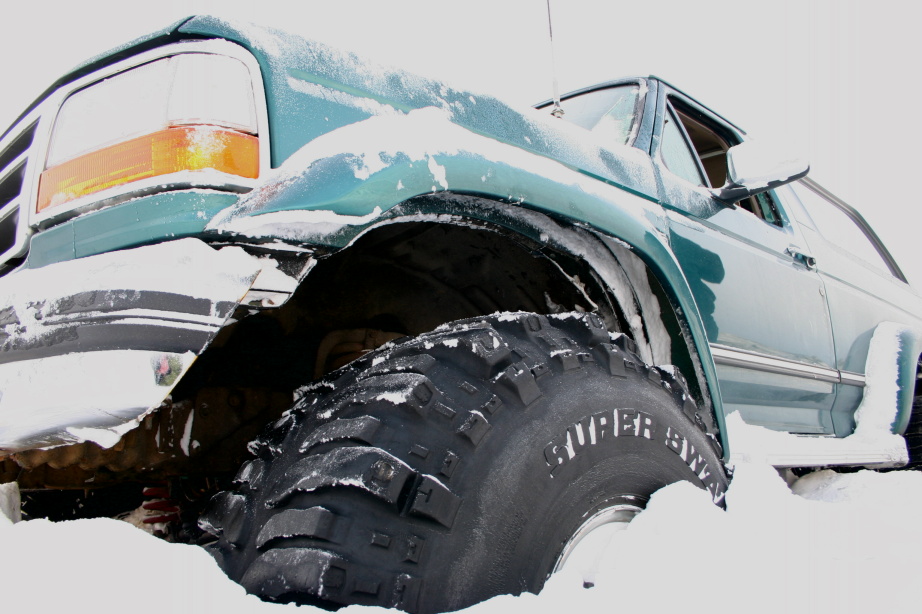 Snow-covered teal Ford Bronco on oversized Super Swamper off-road tires, low front angle