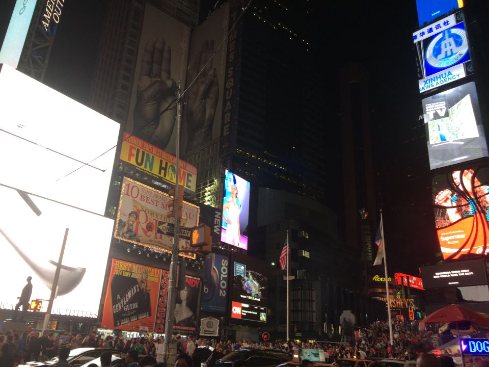 Times Square billboards display advertisements and news at night, with crowds and cars below