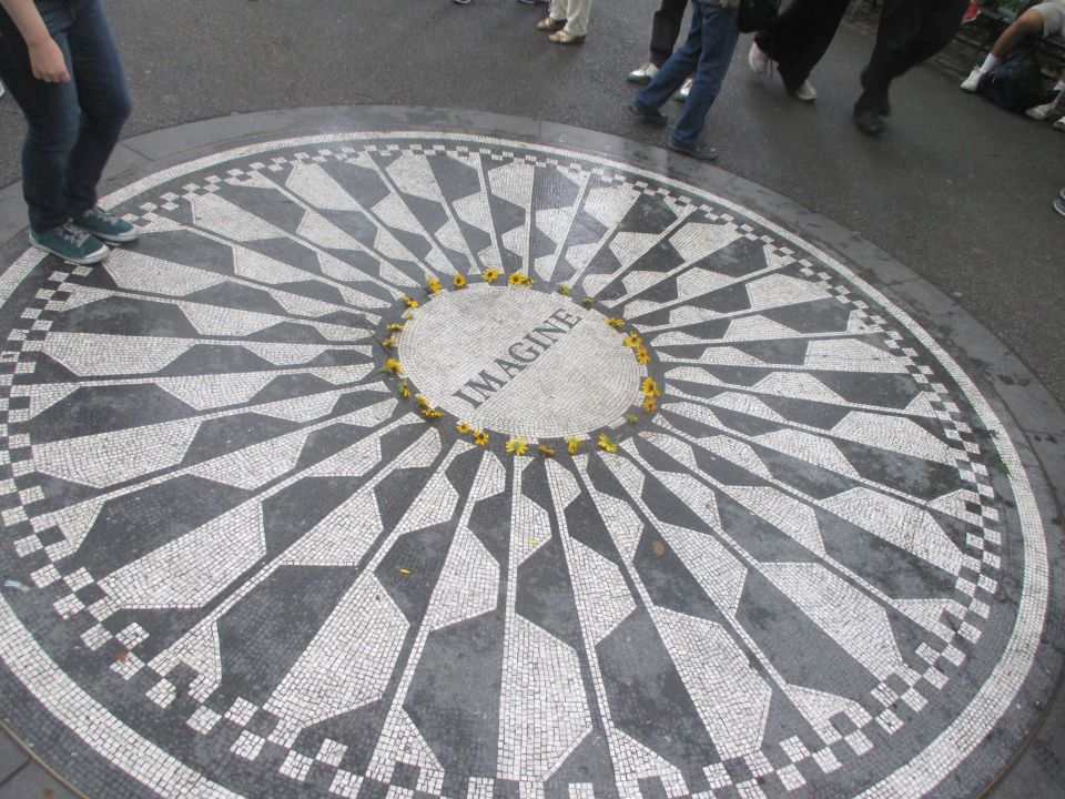Circular mosaic on pavement with the word IMAGINE in the center. People's legs are visible around the edges