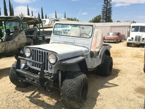 1968 Ford Mustang - A weathered Jeep CJ-5 sits in a junkyard, surrounded by other vehicles under a clear sky.