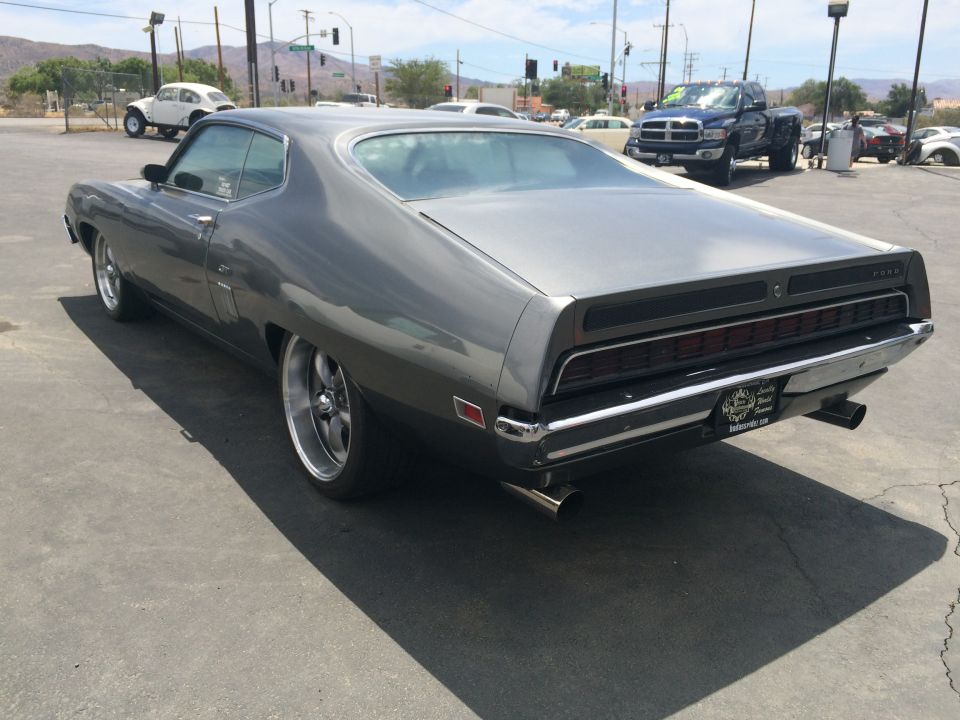 Gray 1970 Ford Torino GT coupe, rear view, parked on asphalt lot with a blue pickup truck and white off-road vehicle in background