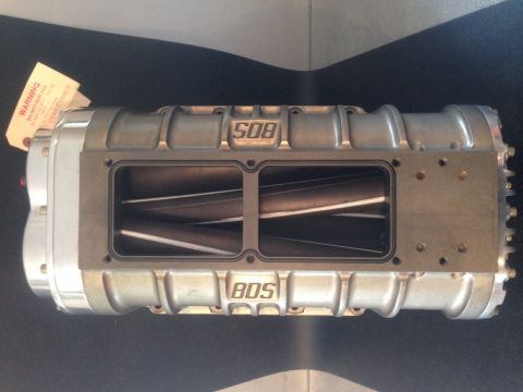 1965 Ford Fairlane - A silver supercharger with visible internal rotors is displayed, with a warning tag attached.