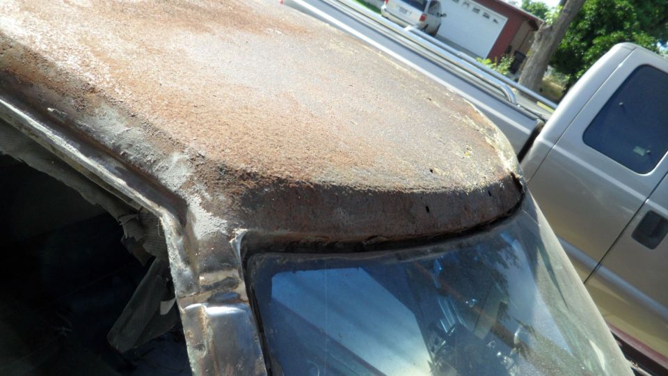 Rusted roofline and windshield frame of a vehicle, with a pickup truck visible behind