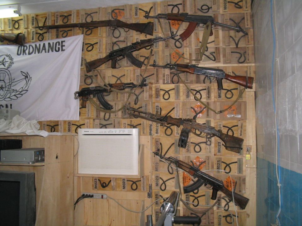 Wall display of several rifles and machine guns mounted on wooden crates. A white flag with the word "ORDNANCE" is on the left