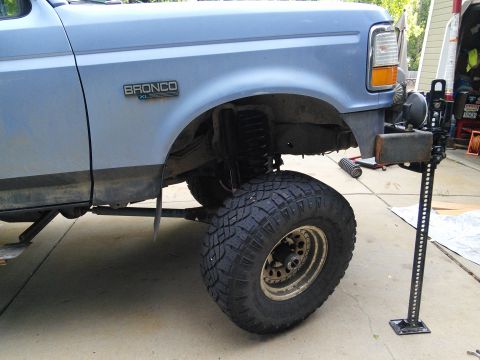 1996 Ford Bronco - A blue Ford Bronco is shown with its front passenger side lifted by a jack, revealing its suspension and large off-road tires.