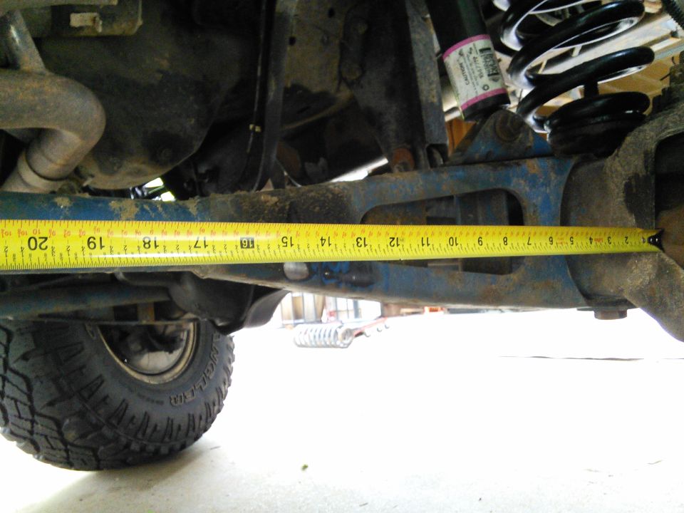 Measuring tape across blue suspension arm on vehicle underside. Coil spring and tire visible