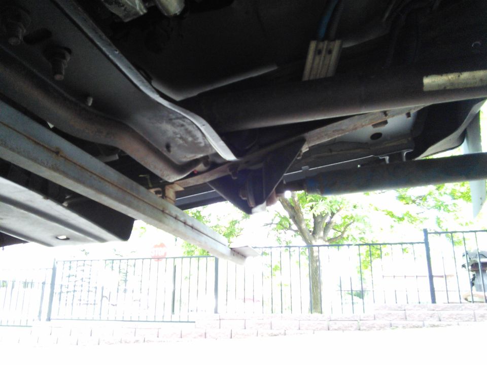 Underside of a vehicle showing frame rails, exhaust pipe, and suspension components. A fence and trees are visible in the background