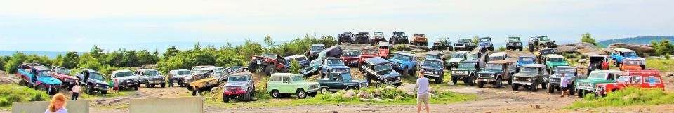 Many off-road vehicles parked on a rocky, grassy hill under a cloudy sky