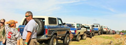 1979 Ford Bronco - A line of modified Ford Broncos and other trucks are parked on a dirt road under a clear blue sky.
