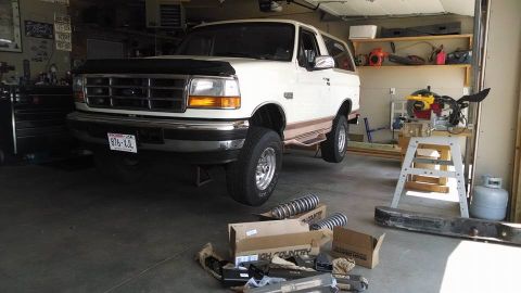 1994 Ford Bronco - A white 1994 Ford Bronco is parked in a garage with new suspension parts laid out, ready for installation.