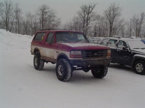 1994 Ford Bronco - A red and white 1994 Ford Bronco with lifted suspension sits in a snowy lot next to other vehicles.