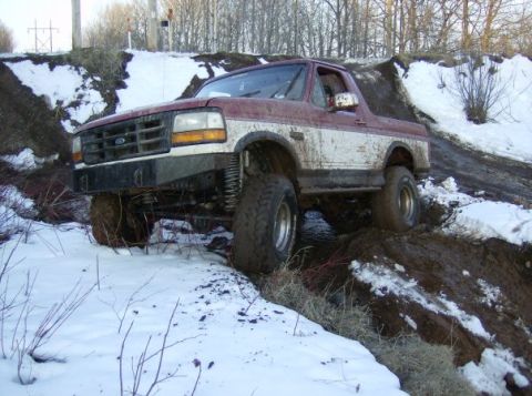 1994 Ford Bronco - Maroon and white Ford Bronco with lifted suspension navigates a challenging muddy and snowy terrain.