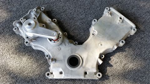 2003 Ford Crown Victoria - reverse side of a cast aluminum V-engine timing cover, showing a center seal bore and many bolt holes, laid on grey carpet.