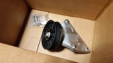 2003 Ford Crown Victoria - black grooved idler pulley mounted on a machined aluminum bracket sits in a cardboard box beside a bag of bolts.