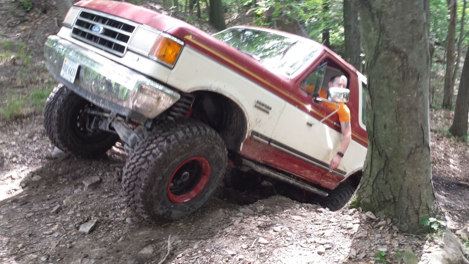 Ford Bronco with lifted suspension and large tires navigating a wooded trail, driver visible