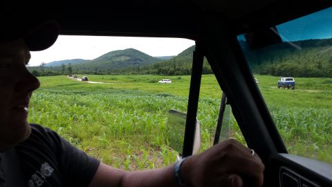 1979 Ford Bronco - A convoy of SUVs and trucks navigates a dirt road through a lush green field.