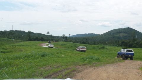 1979 Ford Bronco - Several vehicles, including a blue and white 1979 Ford Bronco, navigate a dirt path through a vast, green, hilly landscape.