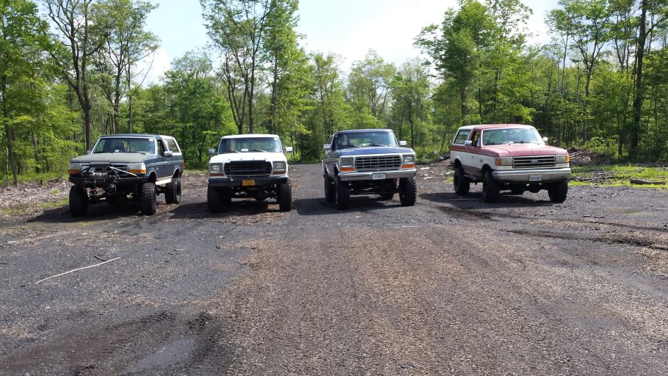 Four Ford Broncos parked on a gravel lot with trees in the background. The Broncos appear to be from the late 1980s