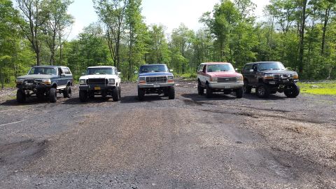 1979 Ford Bronco - A group of four Ford Broncos and a Ford F-150 pickup truck are parked in a gravel lot with trees in the background.